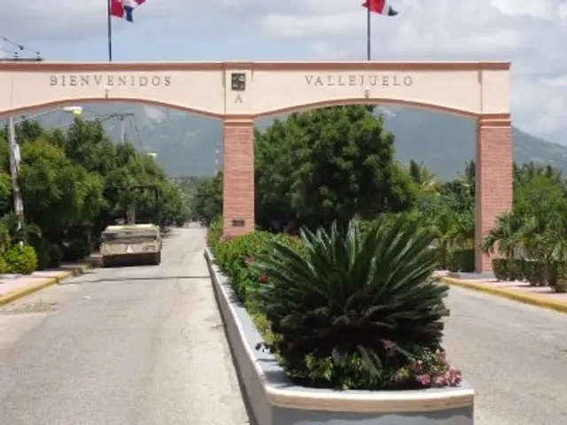 The image shows the entrance to Vallejuelo, Mexico, with an arch with text on it, flags with poles,...