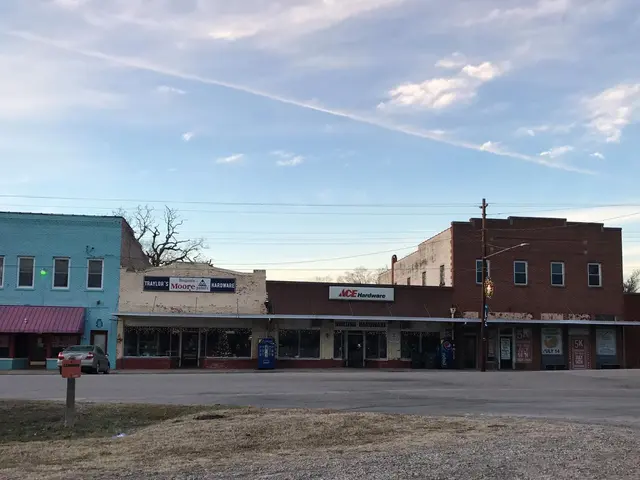 The image shows an empty street in the middle of a small town, with buildings, stores, name boards,...