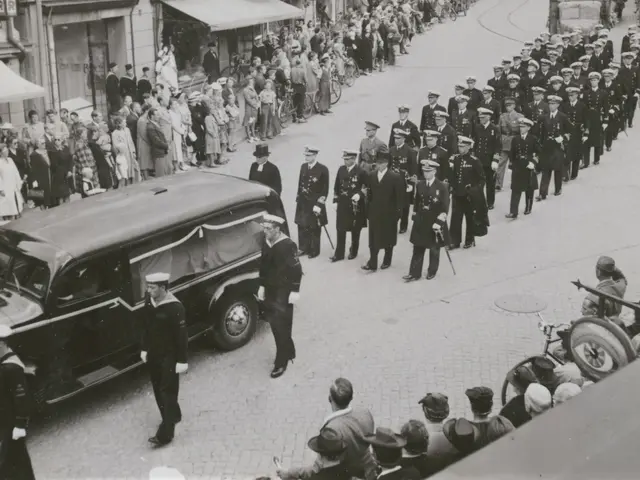 The image shows a black and white scene of a funeral procession with a hearse carrying the body of...