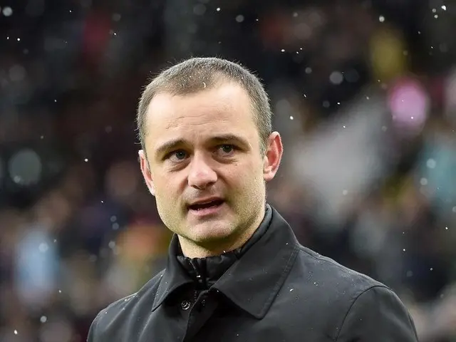 The image shows a man in a black jacket giving a thumbs up, with a blurred background. He is...