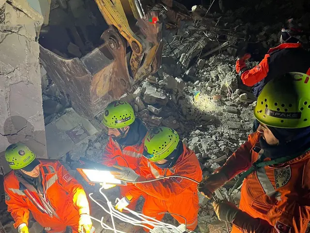 The image shows a group of people in orange uniforms and green helmets working on a pile of rubble,...