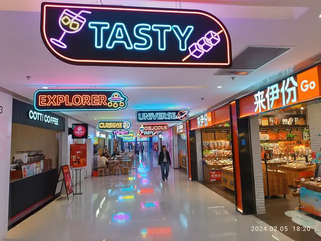The image shows a bustling shopping mall with people walking around, stores with name boards,...