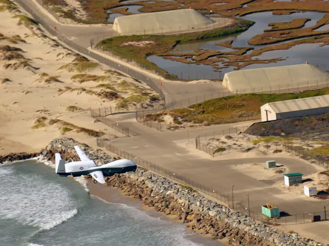 The image shows an RQ-1 Predator Unmanned Aerial Vehicle (UAV) flying over a beach next to a body...