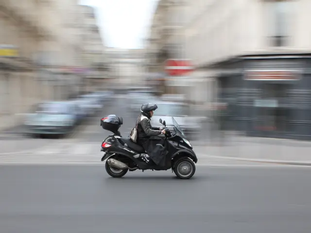 The image shows a person riding a motor scooter down a city street, wearing a black outfit and a...