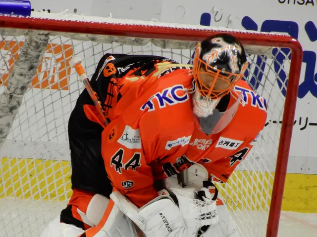 The image shows a hockey player in an orange and white uniform making a save in front of a goal,...