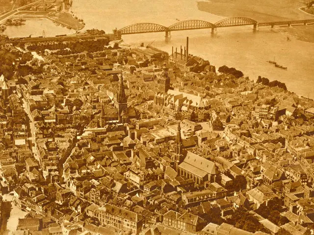 The image shows an old black and white photo of a city with many buildings, trees, and a bridge...