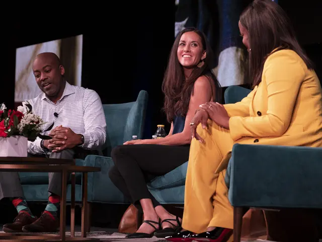 The image shows three people sitting on a couch in front of a screen, with a flower vase and a...