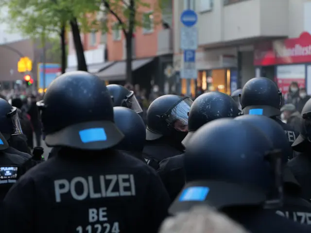 The image shows a group of police officers standing in front of a crowd of people wearing black...