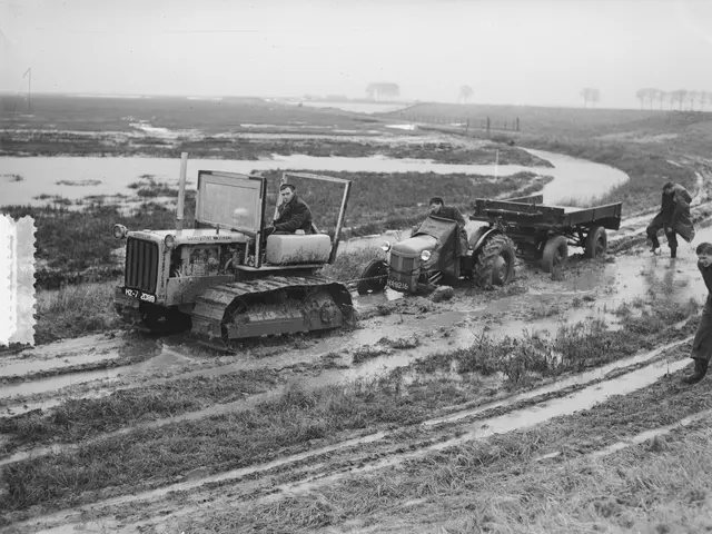 The image shows a black and white photo of a tractor driving down a muddy road, with two people...