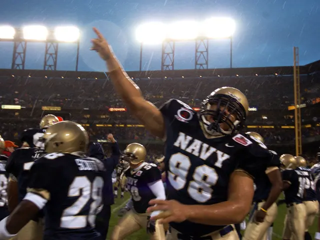 The image shows a group of Navy football players celebrating a touchdown in the rain. They are...