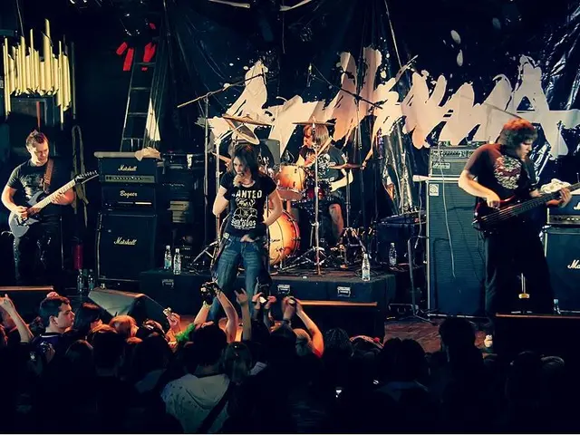 The image shows a group of people on stage playing guitars and drums, with mics and stands set up...