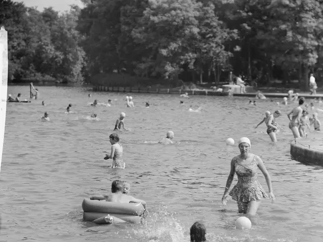 The image shows a black and white photo of people playing in the water at a park. There are trees...