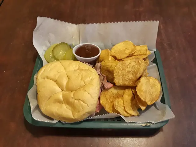 The image shows a tray on a table with a hamburger, chips, and pickles. The hamburger is a classic...