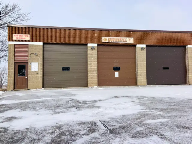 The image shows a fire station with two garages in the middle of a snowy parking lot. The building...