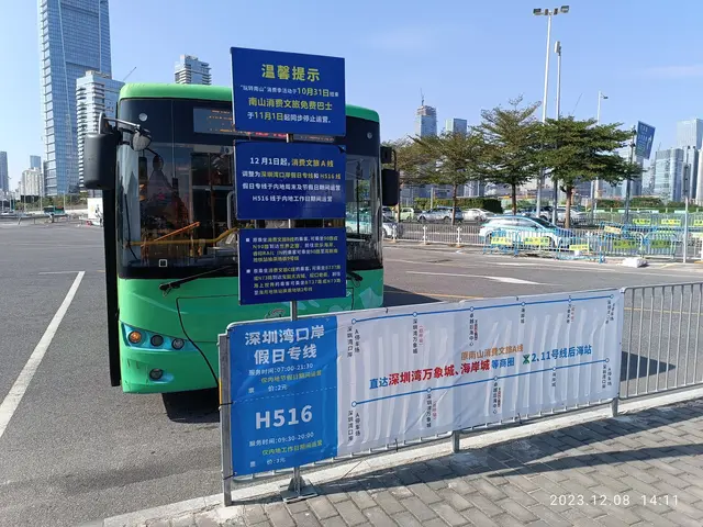 The image shows a green bus parked on the side of a road, surrounded by a metal fence, a banner...