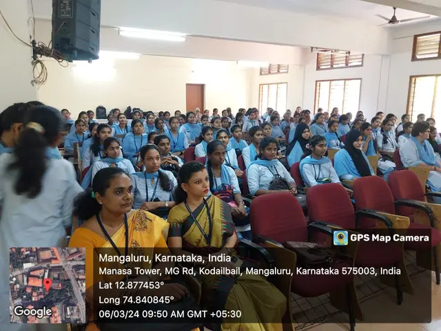 The image shows a group of people sitting in chairs in a room with a wall in the background,...