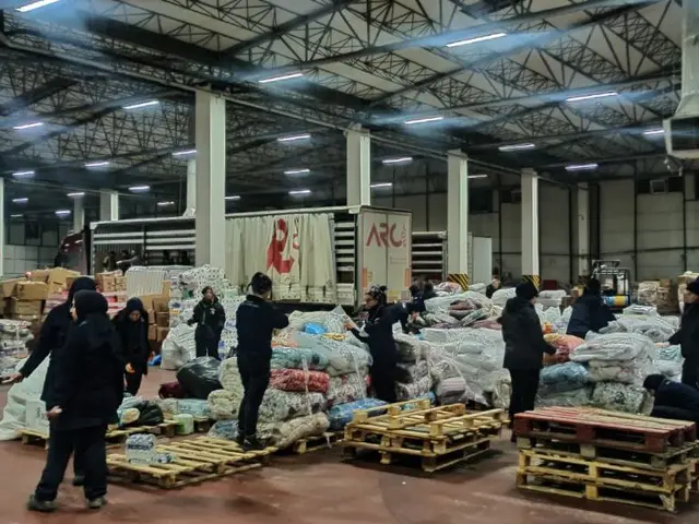 The image shows a group of people standing around a large warehouse filled with bags of food,...