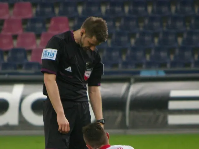 The image shows a soccer player sitting on the ground next to a referee, both wearing sports dress...