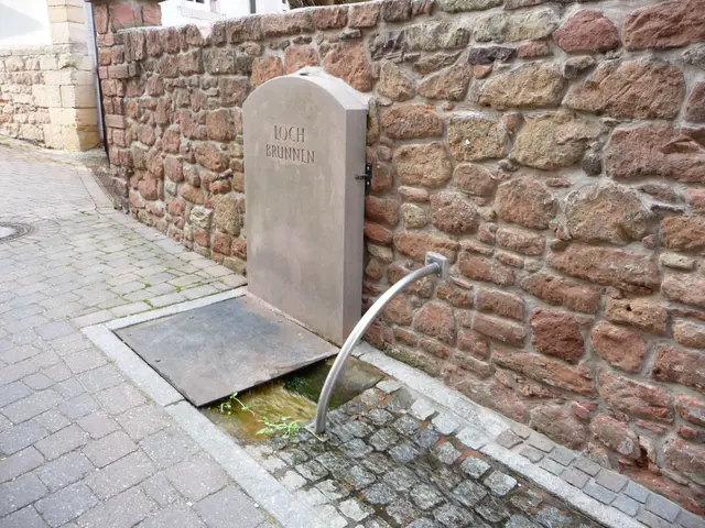 The image shows a stone wall with a metal pipe running along it, a small stream running through it,...