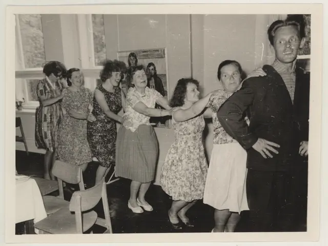 The image shows a group of people dancing in a room with chairs and a table covered with a white...