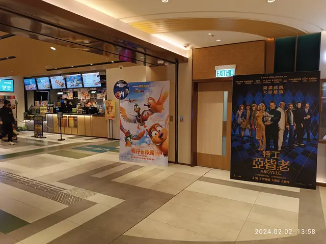 The image shows the lobby of a movie theater with a large poster on the wall, a group of people...
