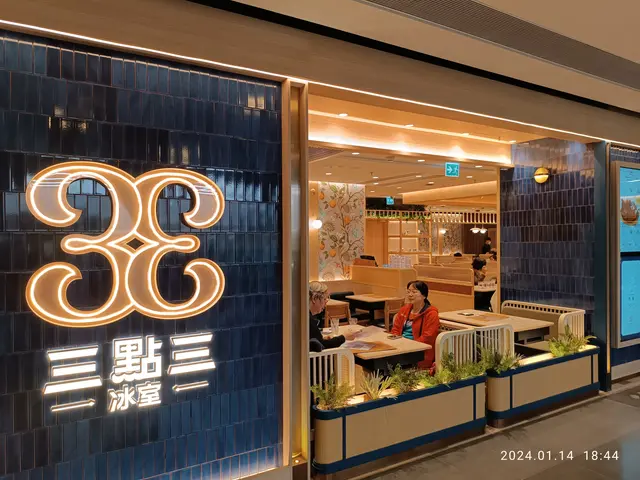 The image shows a restaurant with a neon sign on the side of it. There are people sitting on chairs...