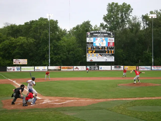 The image shows a baseball game being played on a grassy field with a scoreboard in the background,...
