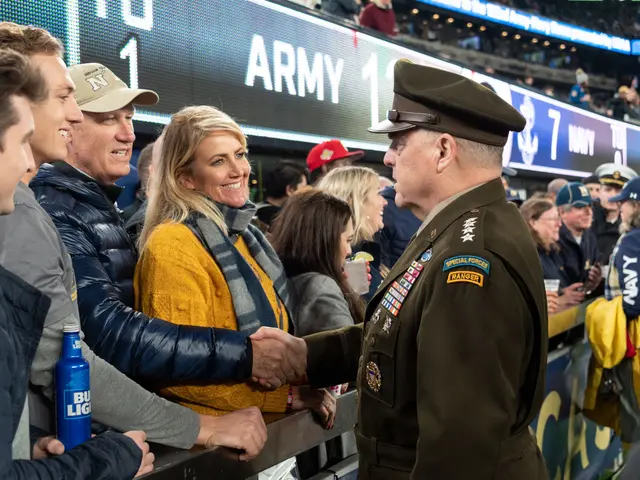 The image shows a man in a military uniform shaking hands with a woman at a football game. There...