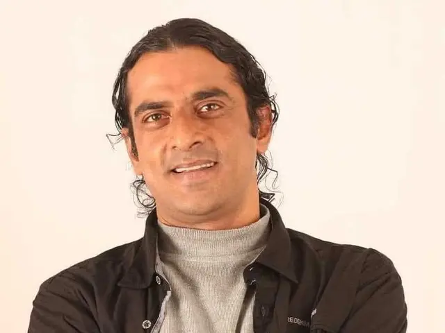 The image shows a man standing with his arms crossed in front of a white background, wearing a...