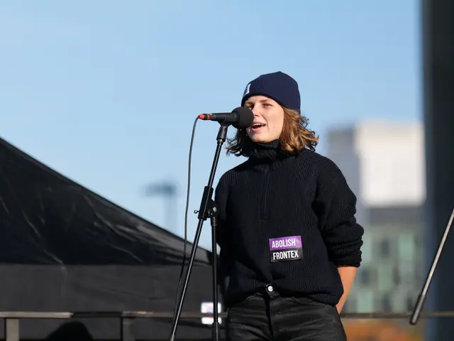 The image shows a woman standing in front of a microphone on a stage, wearing a black dress and a...