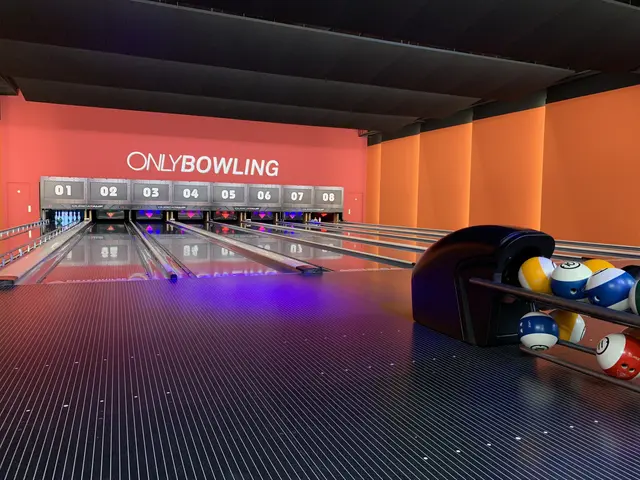 The image shows a bowling alley with a row of bowling balls on the right side, bowling pins set up...