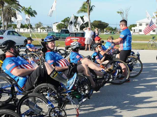 The image shows a group of people in wheelchairs sitting on the side of a road. They are all...