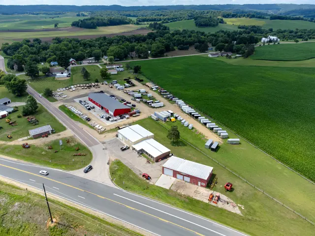 The image shows an aerial view of a farm in the middle of a rural area, with vehicles on the road,...