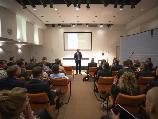 The image shows a group of people sitting in chairs in front of a projector screen, with a man...