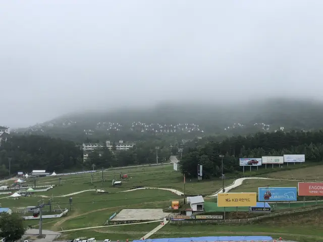 The image shows a view of a ski slope from the top of a hill, with vehicles on the road below,...