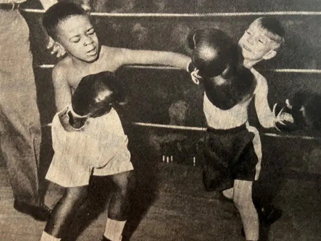 The image shows two young boys boxing in a black and white photo, with a person standing on the...