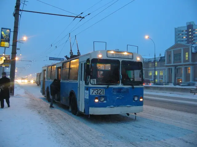 The image shows a blue and white trolleybus driving down a snowy street at night. There are people...
