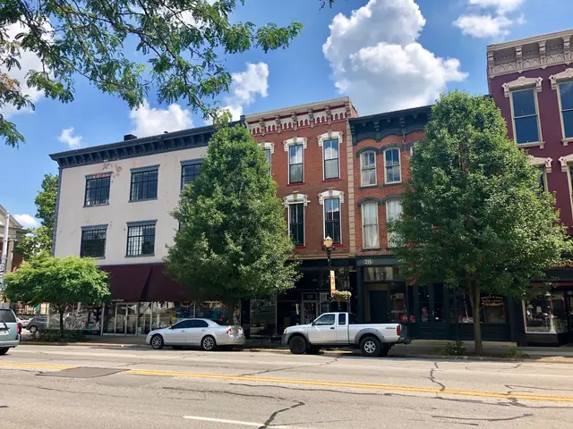 The image shows a city street with cars parked on the side of it, buildings with windows, trees,...