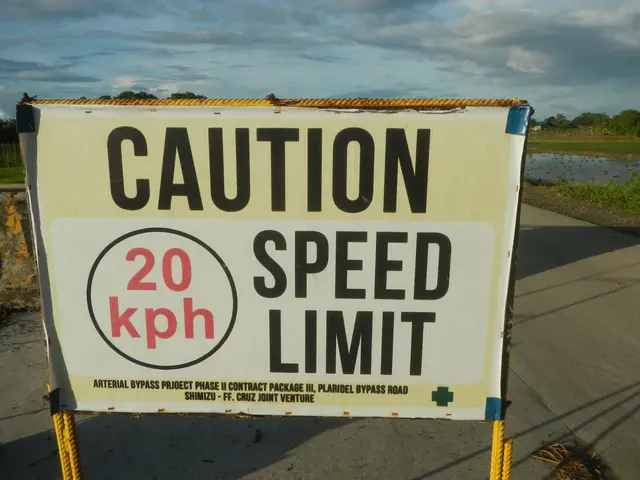 The image shows a caution speed limit sign on the side of a road, surrounded by a wall, grass,...