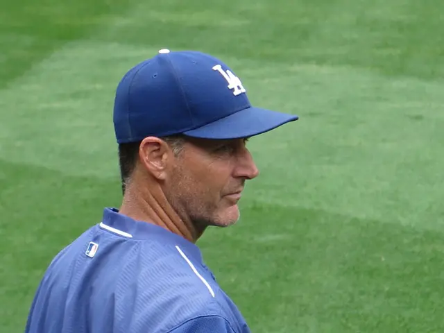 The image shows a man wearing a blue t-shirt and a blue cap standing on top of a baseball field. He...
