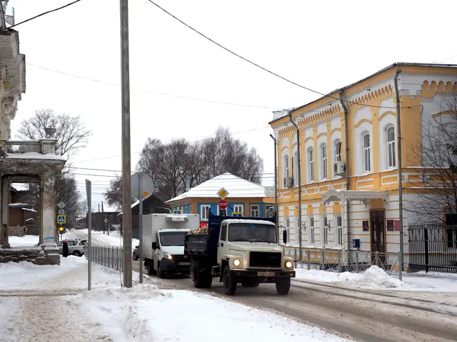 The image shows a truck driving down a snowy street lined with tall buildings. The street is...