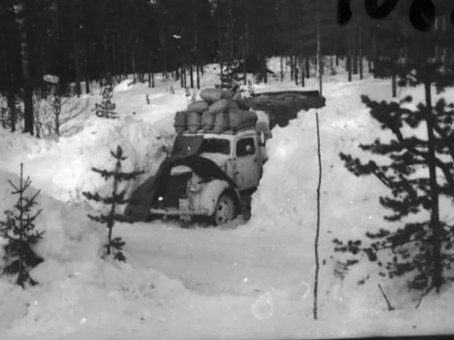 The image shows a black and white photo of a truck stuck in the snow, surrounded by trees. The...