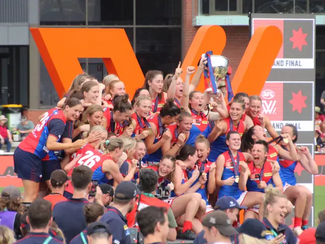 The image shows a group of women's AFL players celebrating with a trophy in front of a crowd. They...