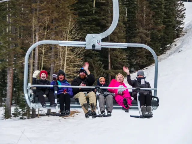 The image shows a group of people riding on top of a ski lift, wearing coats, caps, gloves, and...