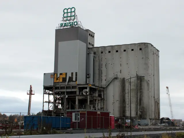 The image shows a large cement building sitting on the side of a road, surrounded by plants, a...