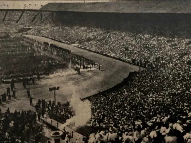 The image shows an old black and white photo of a large crowd of people in a stadium, with a road...
