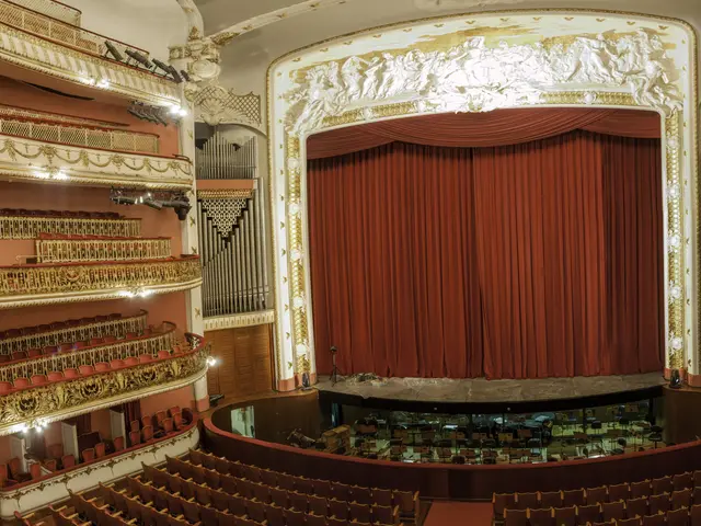 The image shows an inside view of a large auditorium with a red curtain, chairs, railings, lights,...