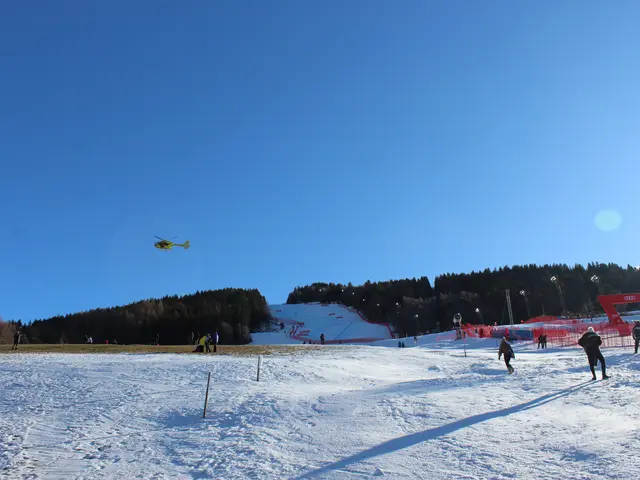 The image shows a group of people skiing down a snow covered slope, surrounded by poles, a fence, a...
