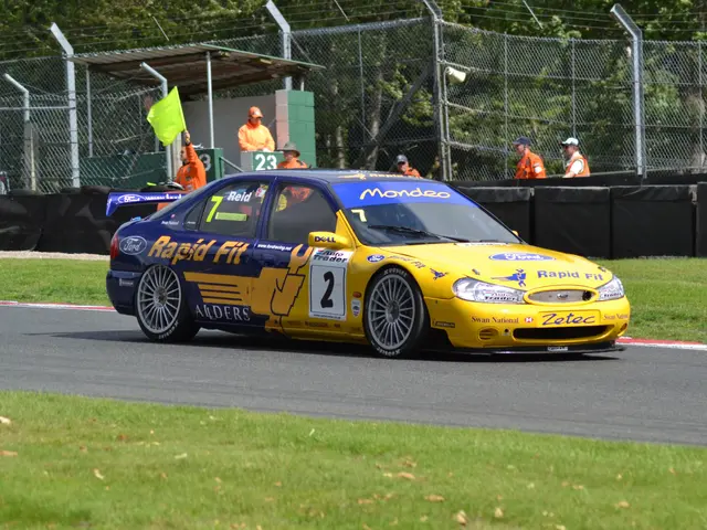 The image shows a yellow car driving down a race track surrounded by grass on either side. In the...