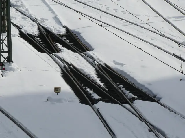The image shows a train track covered in snow next to a building, with electric poles and wires...